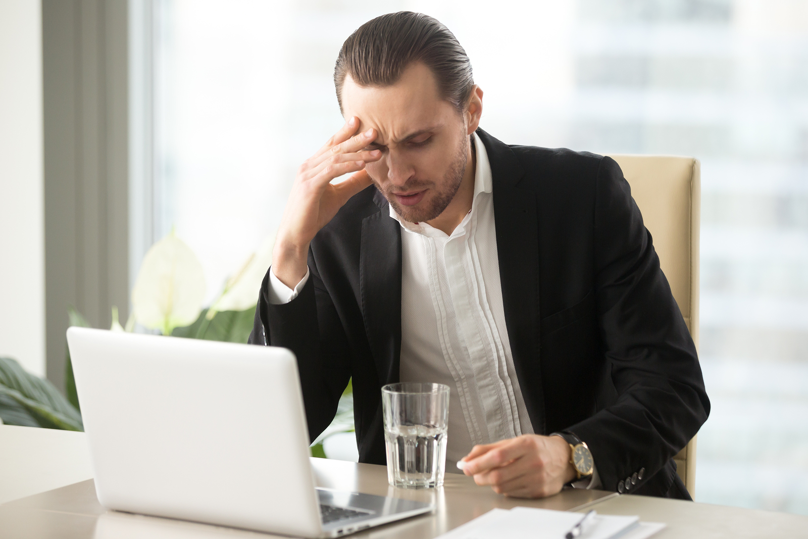 Stressed businessman having severe headache holding painkiller remedy pill Sick CEO with head cold thinking about taking medicine with glass of water Being ill at work stress induced migraine