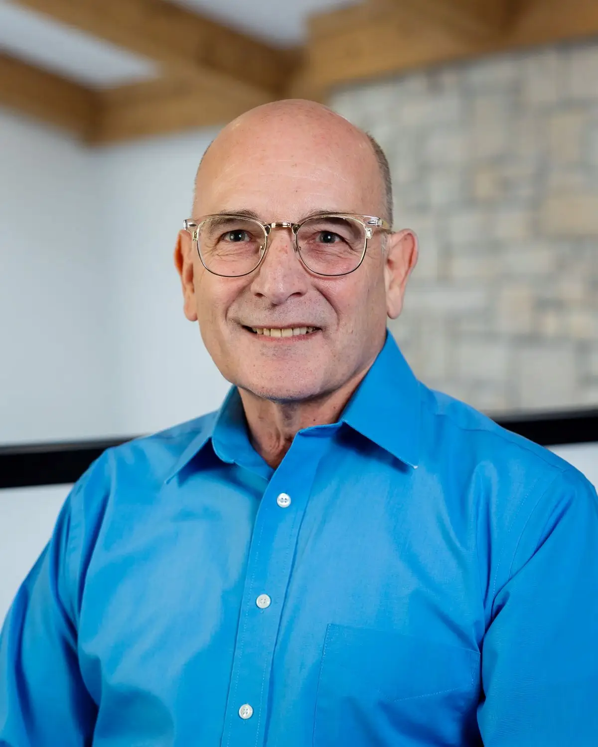 Portrait of an older bald man with glasses wearing a bright blue button down shirt smiling at the camera indoors